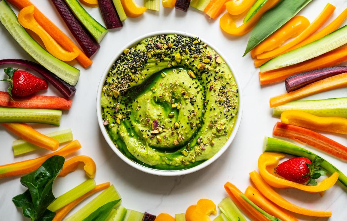 Wild garlic hummus in a bowl topped with pistachios, light and black sesame seeds, and a drizzle of green wild garlic oil; surrounded by vegetable sticks made from carrots, celery, bell peppers, and cucumber