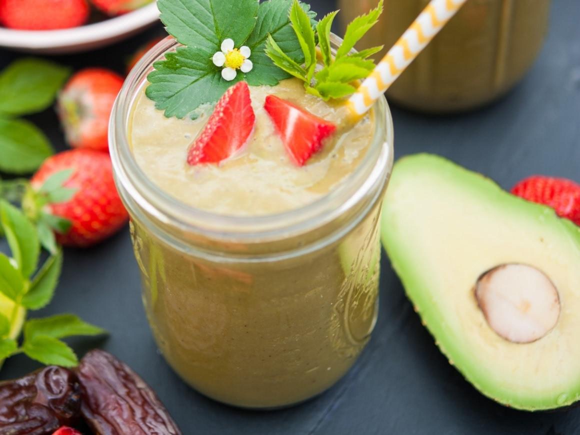 Green smoothie, next to it a sliced avocado and other fruits and leafy greens.